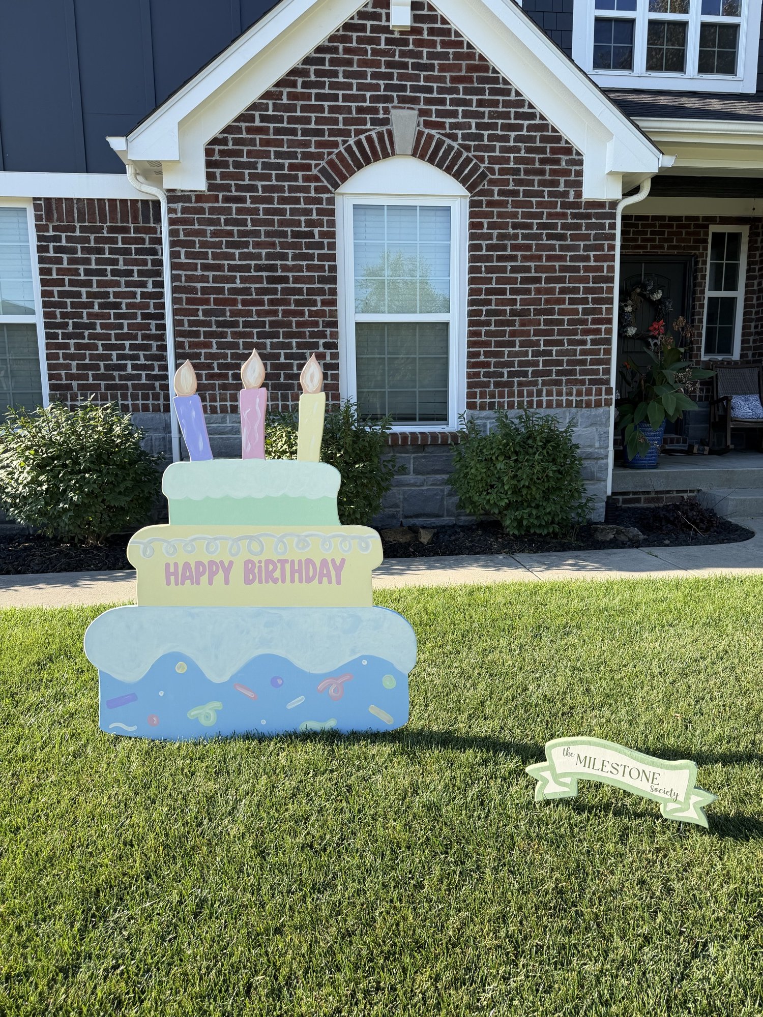 Hand-painted three-tier birthday cake yard sign with pastel candles outside a brick home